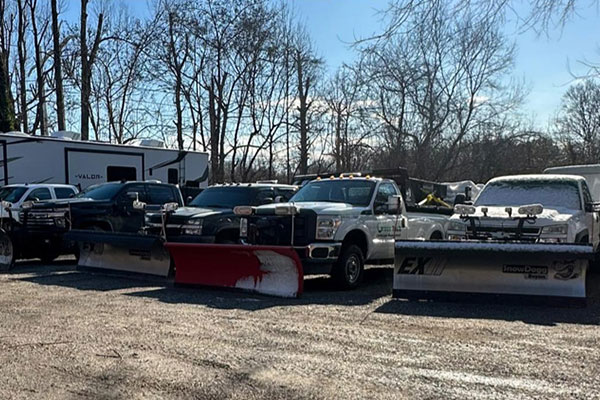 Four snowplow trucks parked in a gravel lot next to a camper, with trees in the background on a clear day.
